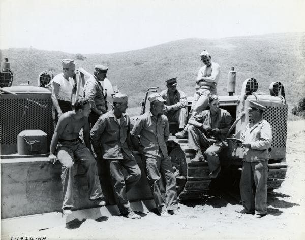 Soldiers sit around International crawler tractors, waiting to receive instruction from a man during a lunch break. The original caption reads: "Group receiving instruction from sergeant in charge and the same group at 'chow' time, hot food being brought up the rough trails by K-3 pickup from mess camp several miles below."