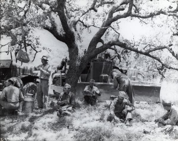 Soldiers sitting on the ground under the shade of a tree to eat lunch. Construction equipment, including an International crawler tractor, is in the background.