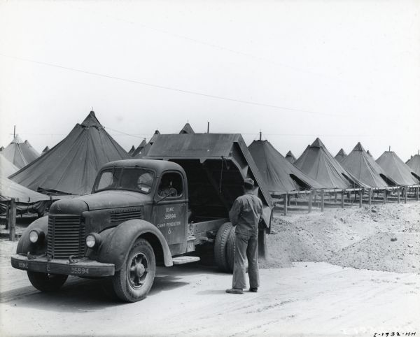 A man stands near an International dump truck marked: "USMC 35894 - Camp Pendleton." The truck bed is raised, releasing a pile of dirt in front of an encampment of tents.