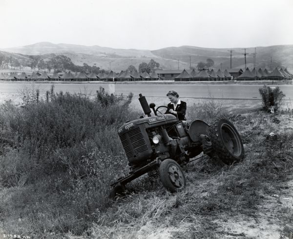 A woman uses an International Model A tractor to mow grass on the side of a hill at Camp Pendleton. In the background is an encampment of tents set up at the base of hills. The original caption reads: "International Model A tractors are used to keep grass and weeds mowed on the Camp Pendleton airfield and women civil service operators employed by the United States Navy Department operate them."