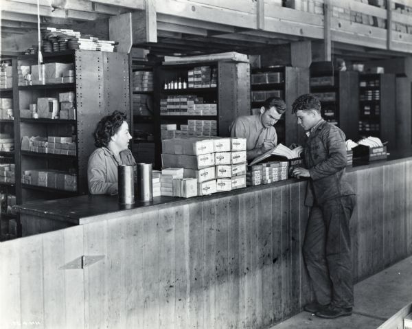 A woman and two men are standing by the counter of Post Motor Transport Department's parts room. Boxes are stacked on the counter; shelves of more boxed supplies are in the background. The original caption reads: "This is a view of the Post Motor Transport Department's parts room, which is 30 x 60 feet in size and has 8 double rows of well-stocked metal bins."