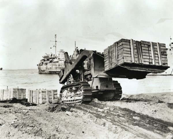 Marines use an International TD-9 crawler tractor (TracTracTor) and dozer shovel loader to unload supplies on a beach. A ship is in the background. The original caption reads: "Towing four thousand pounds of supplies and lifting another two thousand by means of the blade extension and cable, the International TD-9 and dozer shovel loader helps Marines unload supplies at Hawaii."
