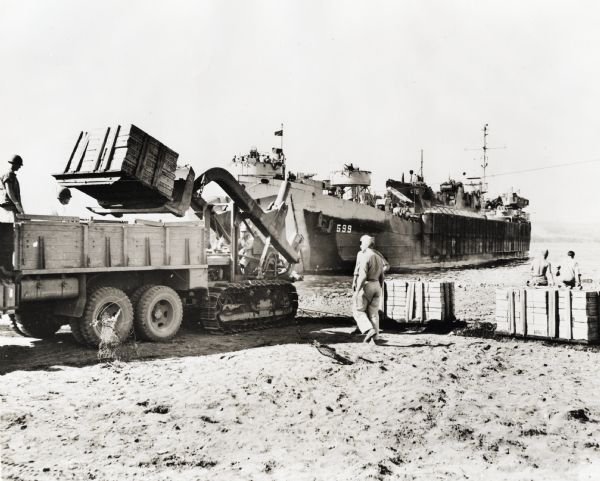 Marines use an International TD-9 crawler tractor (TracTracTor) with a dozer shovel loader to fill a truck parked on a beach. A ship is in the water behind the truck. The original caption reads: "QUICK LOADING----On a beach of a Hawaiian island, Marines employ an International TD-9 with dozer shovel loader to fill a truck. The TD-9 has a two load of four thousand pounds of pallatized supplies."