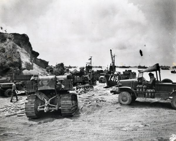 Marines use an International TD-18 Diesel crawler tractor (TracTracTor) to unload supplies and equipment from a ship. Many ships are in the water in the background. The original caption reads: "LANDING OPERATIONS----Supplies and heavy equipment are unloaded ashore on Okinawa Shima on Love Day plus 4. The International TD-18 Diesel TracTracTor in the foreground aids unloading operations."