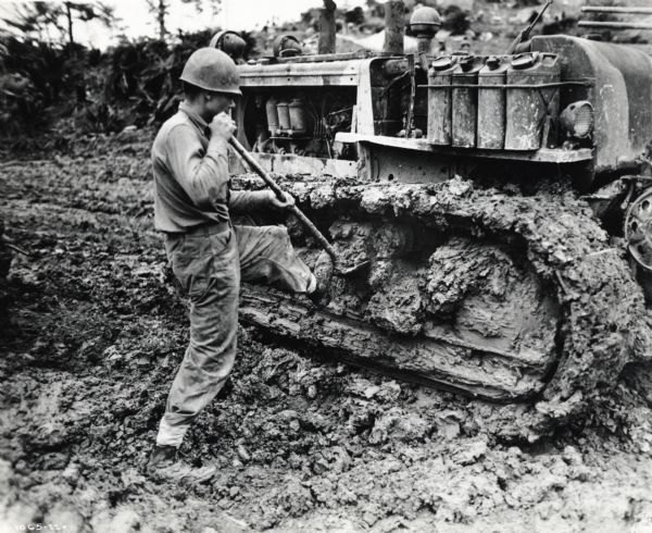 Marine Private First Class Gilbert E. Bailey shovels heavily caked mud from the tracks of his International TD-18 Diesel crawler tractor (TracTracTor). The original caption reads: "WHO'S GOT A TOOTHPICK--At regular intervals, Marine Private First Class Gilbert E. Bailey, 1605 Seventh Avenue, Huntington, W. Va., shovels the Okinawa mud from the tracks of his International TD-18 Diesel TracTracTor in order to maintain a snail's pace along the roads of that Ryukyu Island."