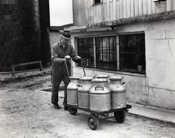 A man pushes a wheeled cart loaded with six milk cans past buildings on International Harvester's Hinsdale experimental farm. The original caption reads: "Pre-production model of McCormick-Deering dairy work on I.H. farm at Hinsdale, Ill."
