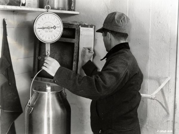 A man inside a building uses a hanging scale to weigh a can of milk. He writes the weight on a chart hanging adjacent to the scale.