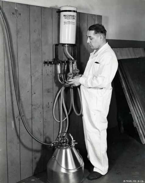 A man holds hoses attached to a wall-mounted McCormick-Deering milker. The original caption reads: "Milker solution rack scene."