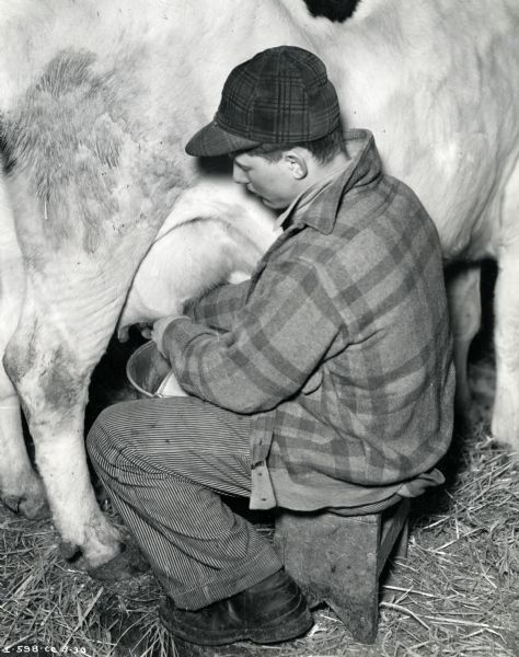 A man sits on a stool to milk a cow by hand. The original caption reads: "Irwin L. Collum, Dousman, Wis. hired man milking for Thomas Ridgeman."