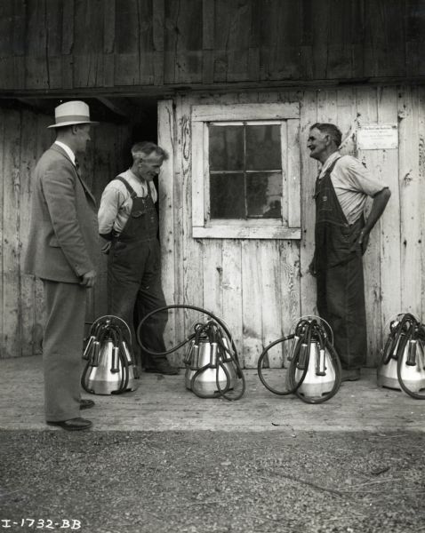 Three men, two wearing overalls and one in a suit and hat, stand outdoors in front of a building beside four McCormick-Deering single-unit milkers on the farm of John Bloomfield.