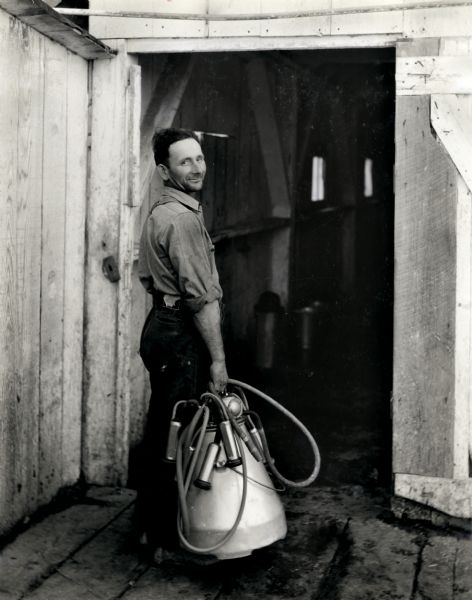 Mr. E. Bognuda stands in a barn doorway while carrying a McCormick milker. The original caption reads: "Milkers on dairy farm owned by Mr. E. Bognuda located in the northern section of California some four miles south of Eureka. Mr. Bognuda says that at present he is milking only thirty-seven cows but generally this number is increased to fifty or fifty-five and he is more than pleased with the performance he has had, and is having, from his International outfit. One photograph clearly reflects this."