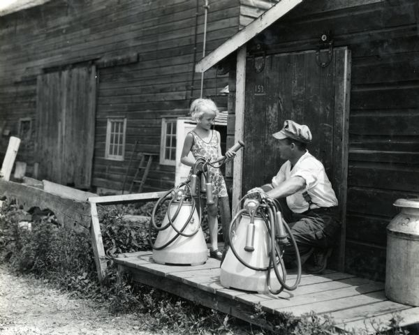 A man crouches beside a McCormick single-unit milker outside a barn, while a young girl standing next to him holds the tubes of another milker in her hands. The milkers are owned by E. Cameron.