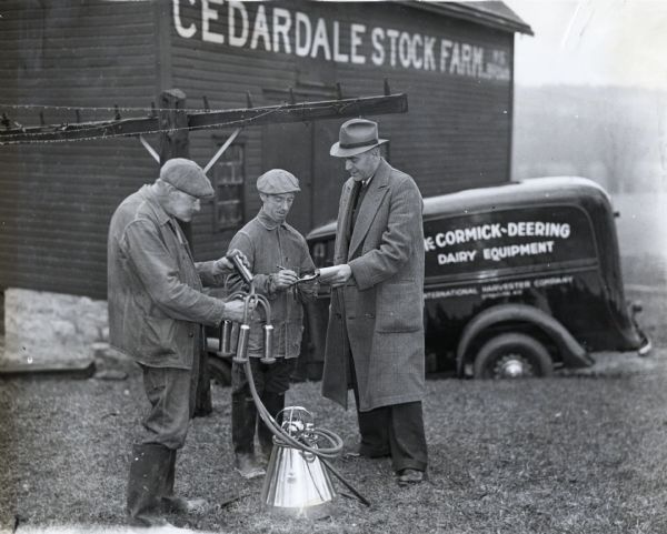 Ralph Lower, dairy equipment salesman from International Harvester's Syracuse branch, sells a farmer a milking machine at Cedardale Stock Farm. A McCormick-Deering truck is in the background next to a barn.