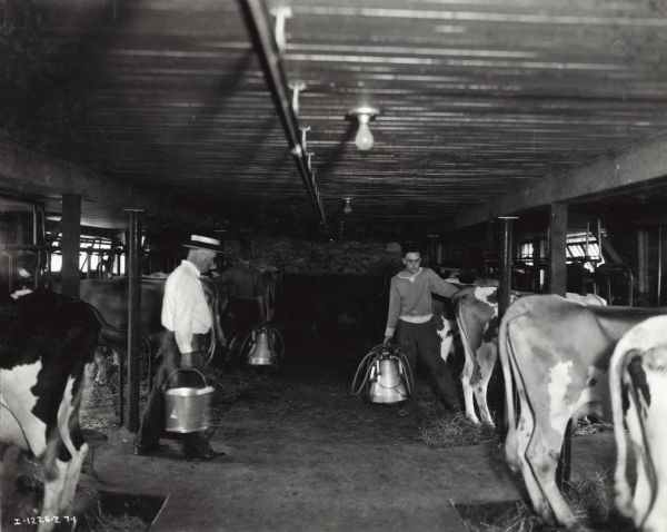 Three men stand between two rows of cows in a barn on the H.C. Slocum farm. The man on the left carries a metal pail, while the man on the right and the man in the background each carry a McCormick-Deering double unit milker.