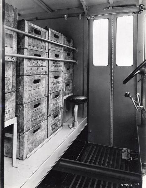 Interior view of the International Model M-3 truck looking toward the steering column and side door. Wooden crates labeled: "Purity Dairy, Springfield Dairy" are stacked behind the driver's seat.