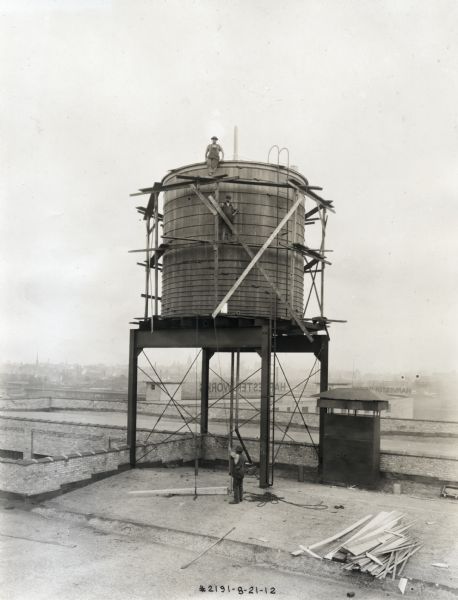 Two men pose on a water tank on top of Building #43 at International Harvester's Milwaukee Works factory, while another man hoists building materials from the roof below. Two large signs reading: "Harvester Works" are near the roof line in the background.