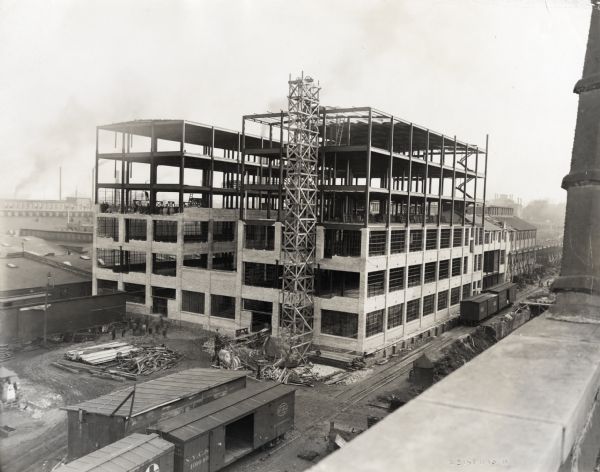 Construction crews erecting a six-story machine shop at International Harvester's Milwaukee Works factory. Men are working on the ground and on an exposed floor of the building. Scaffolding runs the length of the building near railroad cars parked on railroad tracks which run along the side of the building.