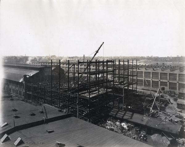 Elevated view of construction of building number 43 at International Harvester's Milwaukee Works. The factory building housed a six story machine shop. Men are using a crane to erect the building's steel frame. Dwellings and church buildings are in the far background.