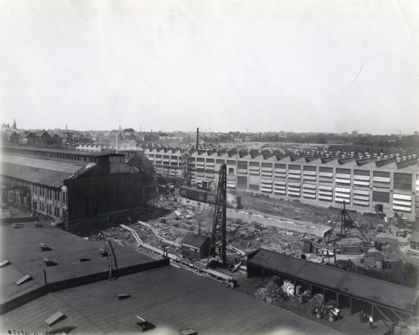 Elevated view of a cleared section of land for a six-story machine shop at International Harvester's Milwaukee Works factory. Other industrial buildings and railroad cars and tracks surround the cleared site, and dwellings and church buildings are in the far background.