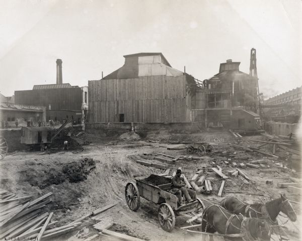 A man uses a horse-drawn wagon to transport building materials on a construction site at International Harvester's Milwaukee Works factory. Men are clearing ground to make space for the six-story machine shop.