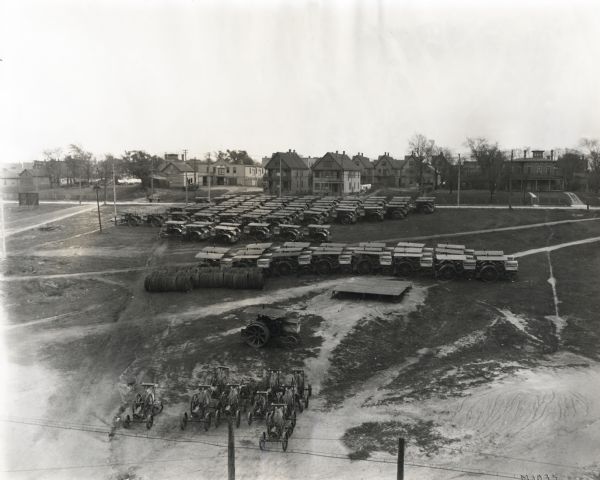Elevated view of tractors awaiting shipment parked in a grassy lot at International Harvester's Milwaukee Works factory. There are houses and commercial buildings in the background.