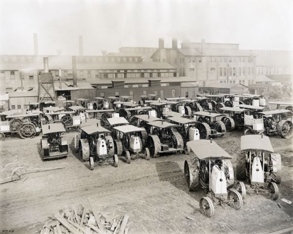 Elevated view of tractors parked in rows in a dirt lot at International Harvester's Milwaukee Works factory. Factory buildings are in the background.