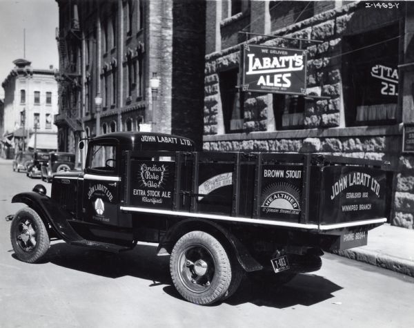 International Harvester truck parked on a city street, possibly in Winnipeg, Manitoba, Canada. The truck was used by John Labatt Ltd. to deliver beer. The text on the truck advertises India pale ale and brown stout. The stone building in the background has a sign reading: "We Deliver Labatt's Ales."