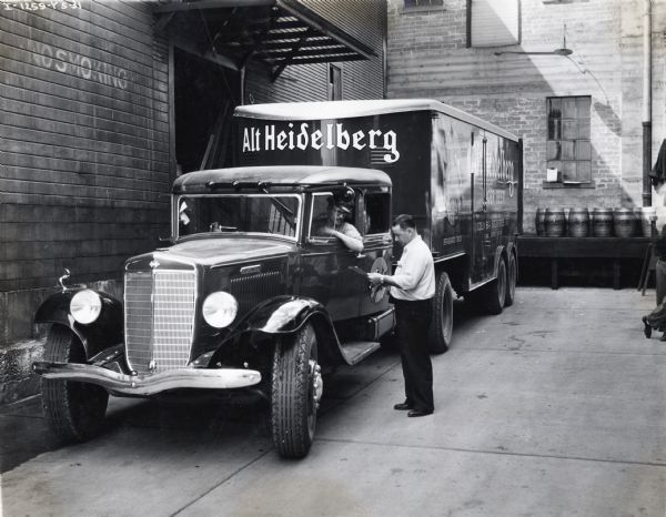 A man holding a clipboard is standing and talking to a driver seated behind the wheel of an International C-60 truck parked in a loading area. The refrigerated truck was used by Columbia Breweries, Inc., of Tacoma, Washington to transport "Alt Heidelberg" beer and had a 14 ton capacity.