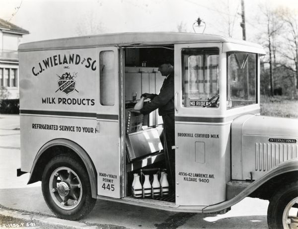 View through the side door of an International milk truck showing a man loading bottles from a refrigerated compartment into a wooden crate. The truck is parked near the curb in a residential area. The truck was used by C.J. Wieland and Son, Inc. to deliver dairy products.