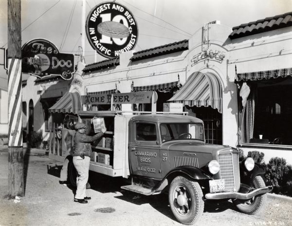 A man handles boxes of beer on the side of an International C-35 truck used by the Cammarano Brothers company. The truck has a wooden sign over the back reading: "Acme Beer, America's Finest," and is parked in front of a restaurant. The original caption reads: "A C-35 owned by Cammarano Bros. of Tacoma, Wash., and used in the distribution of their bottled products, especially in handling beer that is distributed to the many inns and roadhouses in this territory. One picture shows this unit making one of these deliveries and the other taking on its load at the brewery."