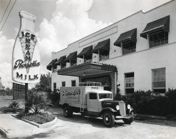 An International C-35 truck used by Poinsettia Dairy Products, Inc., parked outside a company building. A sign in the shape of a milk bottle advertises ice cream and milk near the driveway on the left. The lamps flanking the entrance are shaped like milk bottles and have a "Poinsettia" label.