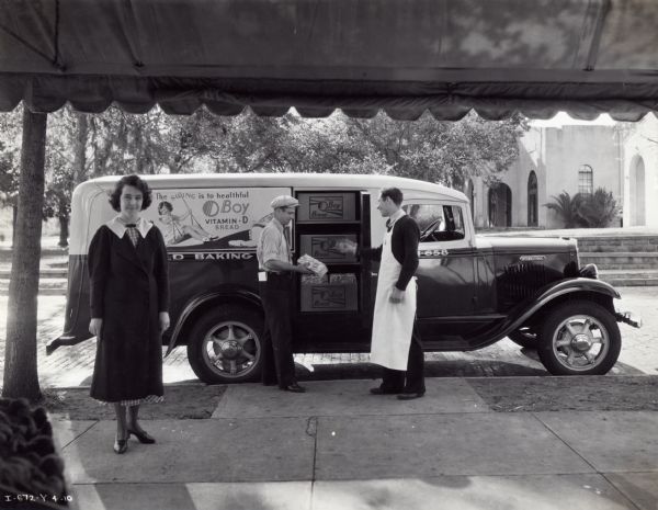 View from under an awning of a man standing next to an International Model C-20 truck used by the Seybold Baking Company. He is handing a loaf of "O Boy" brand bread to a man wearing an apron. The truck features a panel body with a special side door. A woman stands to the left facing the camera. The advertisement on the side of the truck reads: "The swing is to healthful O Boy Vitamin-D Bread."