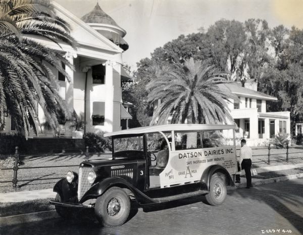 A man unloads milk bottles from the back of an International Model C-1 truck used by Datson Dairies Inc. The truck is parked on a residential street in front of several houses.