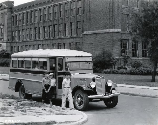 Three boys exit an International school bus in front of what appears to be a school building. Three children sit inside the bus looking out the windows.