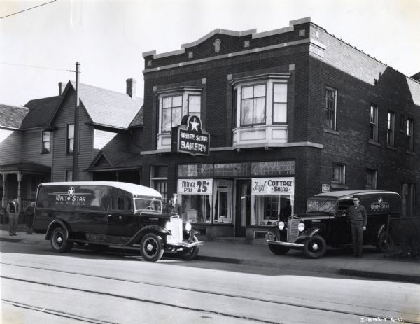Two International trucks used by the White Star Bakery are parked outside the bakery's storefront at 1473 Main Street. Each truck's driver stands beside his vehicle. The original caption reads: "C-1 & C-30 - 157" W.B. Truck - 7' Panel and type AA Panel."