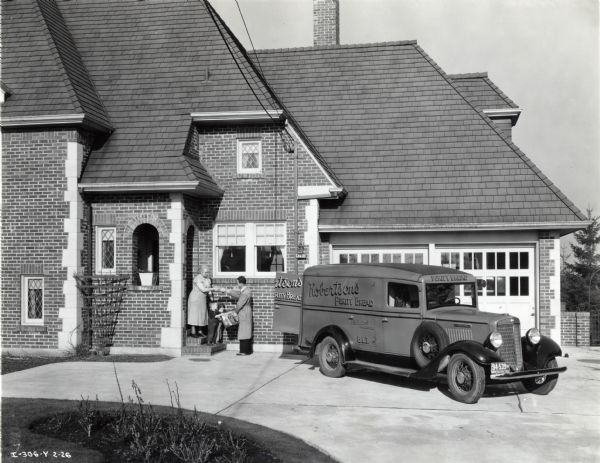 An International truck used by Robertson's Bakery is parked in the driveway of a residence as the truck's driver delivers bread to a woman and two children standing on the doorstep. The text on the truck reads: "Robertson's Purity Bread."
