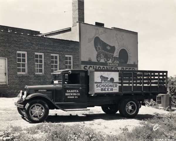 Side view of an International truck used by the Dakota Brewing Company of Huron, South Dakota, parked in front of a brewery building. Both the advertisement on the bed of the truck and the painted sign on the brewery read: "Ask For Dakota Schooner Beer."