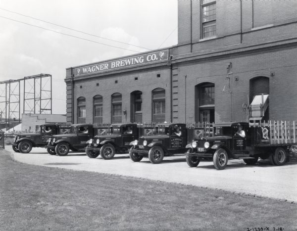 Drivers sit behind the wheels of International trucks owned by Wagner Brewing Company. The trucks are parked in a row outside a brewery building with a sign that reads: "Wagner Brewing Co."
