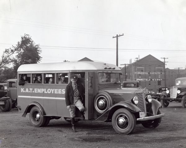 A driver exits an International bus marked: "K.A.T. Employees" in a dirt parking lot. Other passengers sit inside the vehicle, and International's Springfield Works buildings are in the background.