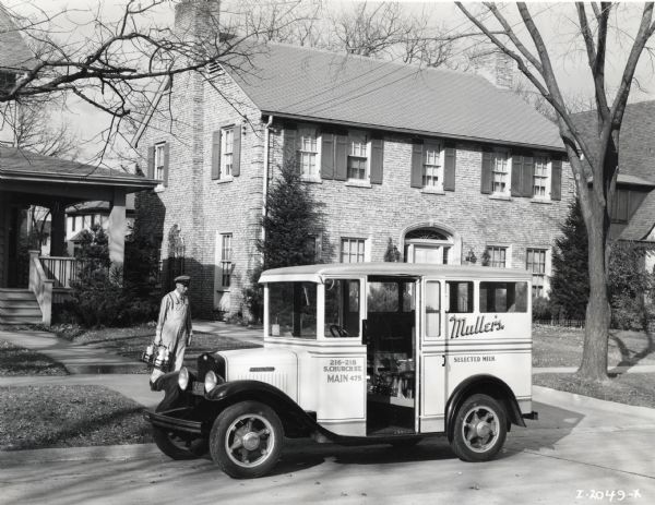 A man carrying a crate of milk bottles and wearing a hat and overalls walks toward an International truck owned by Muller's Union Dairy. The truck is parked by the side of the road in a residential area.