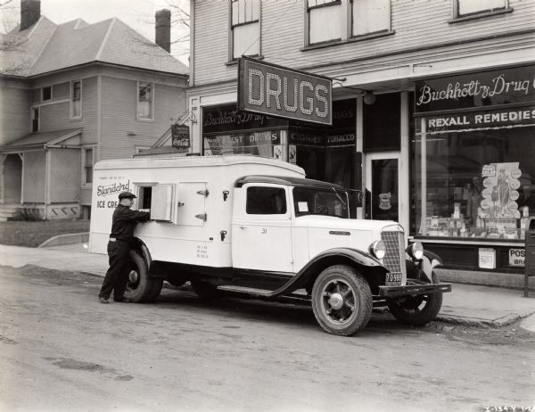 A man reaches into an open door on the left side of a refrigerated compartment of an International truck owned by Standard Ice Cream. The truck is parked in front of Buchholtz Drug Store.