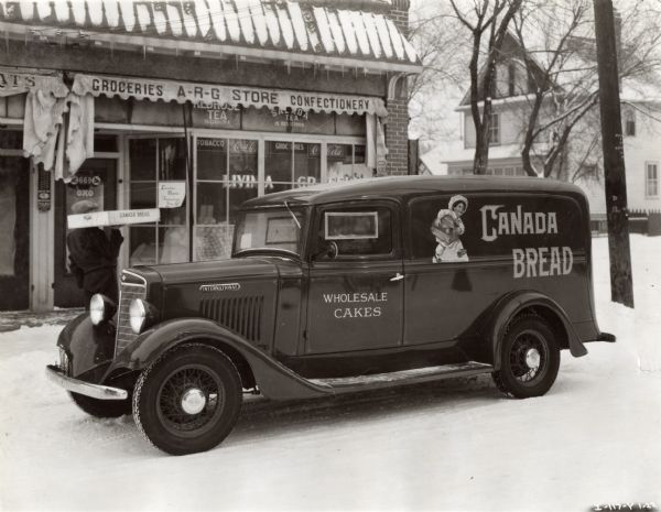 A man unloads a box from an International Model C-1 truck with a 125-inch wheelbase parked in front of a grocery store. The text on the truck reads: "Canada Bread" and "Wholesale Cakes." The store's awning reads: "Groceries A-R-G Store Confectionery."
