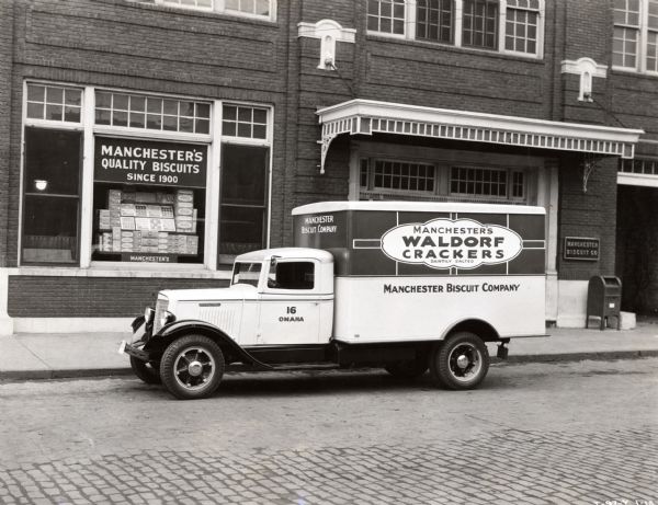 International Model C-30 truck owned by the Manchester Biscuit Company parked along the curb in front of the company's building. A display of biscuit boxes is visible in the building's window under a sign reading: "Manchester's Quality Biscuits / Since 1900."