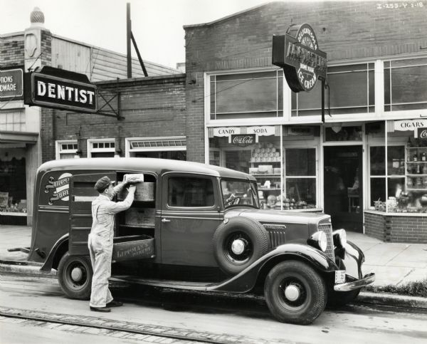 View from street of a man wearing overalls and a hat loading a loaf of Lippman's bread into a box shelved in the back of an International truck owned by Lippman's Bakery. The truck is parked along the curb of a commercial street in front of a hardware store, dentist office, and a delicatessen.