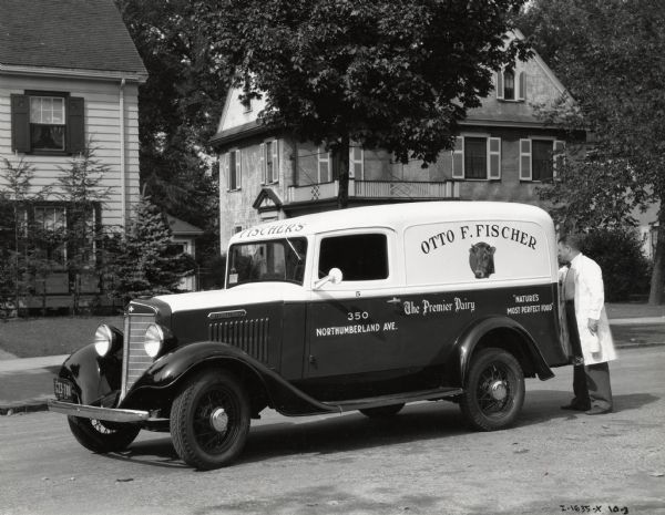 A man wearing a long white coat and holding a bottle of milk is standing near the back of an International truck owned by Fischer's Dairy. The truck is parked in the street in a residential area, and the text on its side reads: "Otto F. Fischer; The Premier Dairy; 'Nature's Most Perfect Food', and 350 Northumberland Ave."