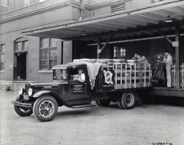 Men standing on a loading dock are placing beer barrels from the Wagner Brewing Company onto the bed of an International truck. A man is sitting in the driver's seat of the truck. The text on the driver's door reads: "L.F. Kruger, Distributor, St. Louis."