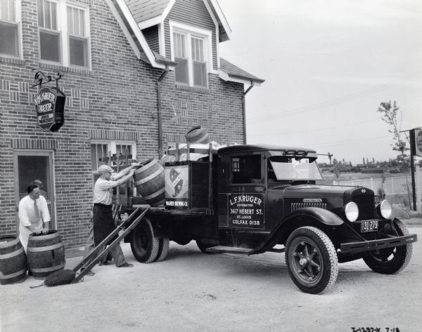 Men loading barrels from Wagner Brewing Company onto the bed of an International truck. They are standing near a doorway to a brick building with an electric sign above their heads that reads: "Wagner Beer / John Munzert. "The text on the passenger-side door reads: "L.F. Kruger, Distributor, 3617 Hebert St., St. Louis." In the far background on the right are houses and trees.