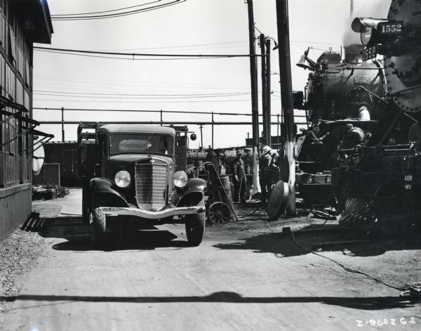 An International Model C-35 is parked near a building. To the right men are working on a locomotive for the Missouri Pacific Railroad. The front of another train engine is in the foreground on the right.