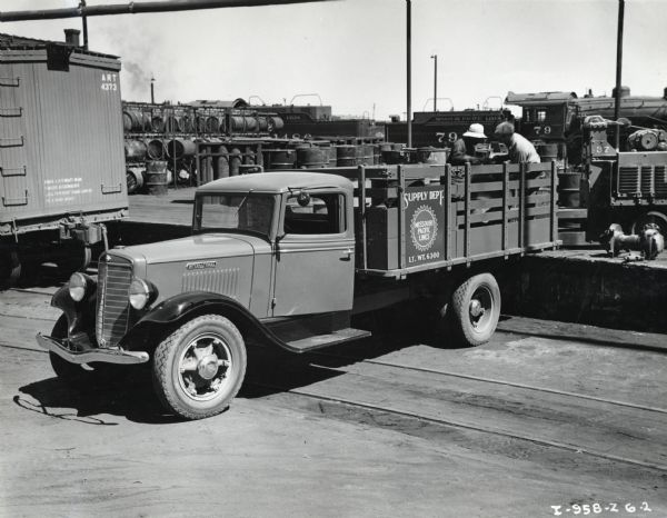 Slightly elevated view of men loading the bed of an International Model C-35 truck parked behind a railroad car. The men are standing on a platform at the Missouri Pacific Railroad yard. Railroad cars, industrial barrels, equipment, and parts are in the background.