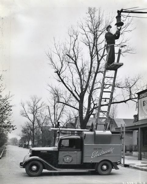 Man standing on a ladder replacing a light on a residential street. The ladder is extended from the bed of an International truck owned by the Indianapolis Power and Light Company. There is a sign on a storefront on the right, and dwellings line the street.